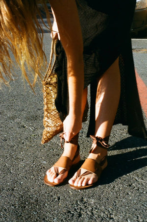 Close up of woman tying Cabo vegan lace up sandals with brown straps styled with black summer dress outdoors