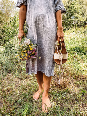Woman holding vegan lace up sandals with neutral ribbons while walking barefoot in nature with flowers, sustainable lifestyle