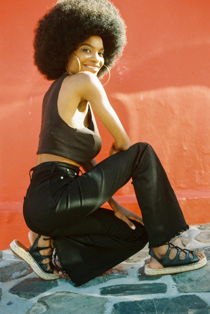 Woman with natural afro hairstyle wearing black top, flared pants, and Rarámuri lace-up espadrille sandals, posing against a bright red wall.