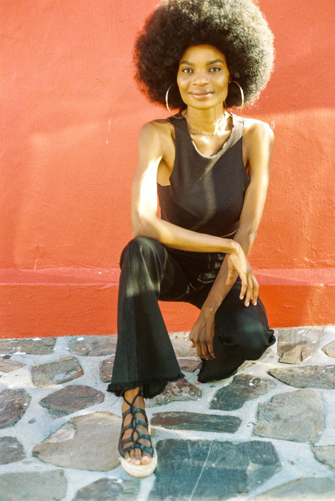 Woman with natural afro hairstyle wearing black top, flared pants, and Rarámuri lace-up espadrille sandals, posing against a bright red wall.