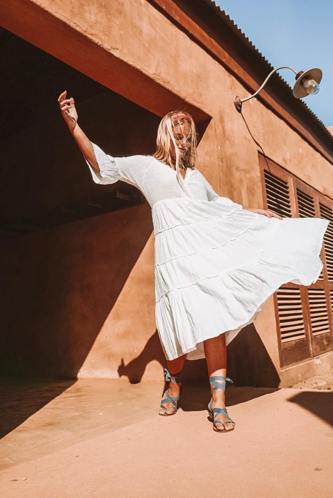 The same woman twirls joyfully in the sunlight, wearing the same dress and blue ribbon sandals against a warm terracotta wall.