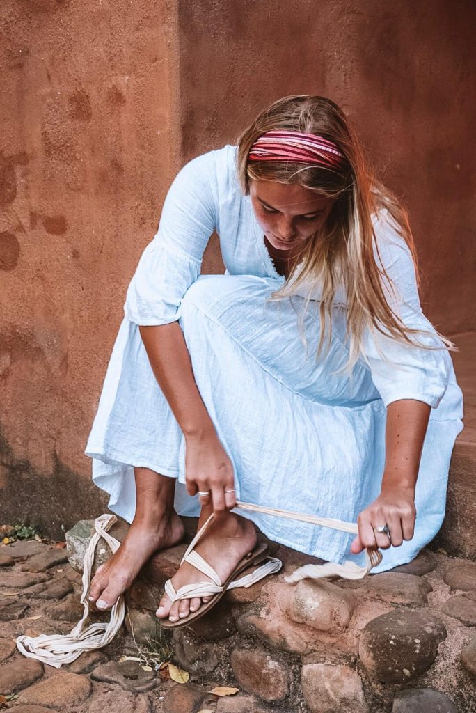 A woman in a light blue flowy dress sits on a stone step, tying the white ribbons of her Rarámuri sandals.