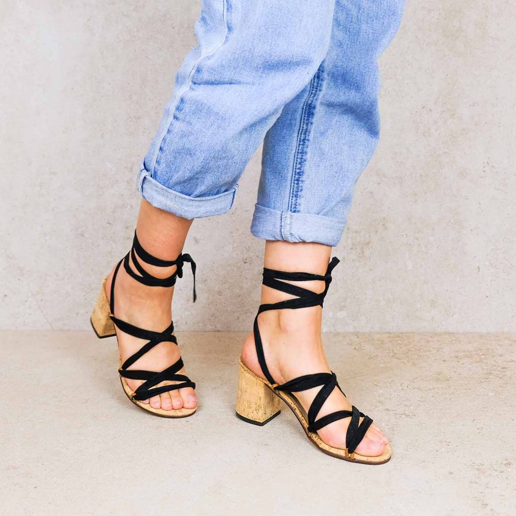 Close-up of legs wearing Rarámuri black ribbon sandals with cork block heels, styled with rolled-up blue jeans.