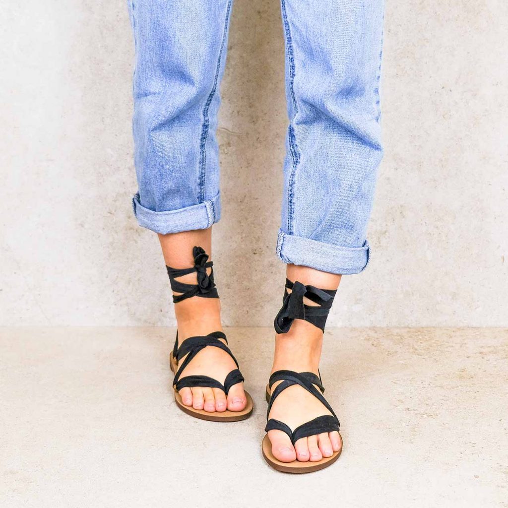 Close-up of feet in Rarámuri flat sandals with black ribbon ties, styled with rolled-up blue jeans.