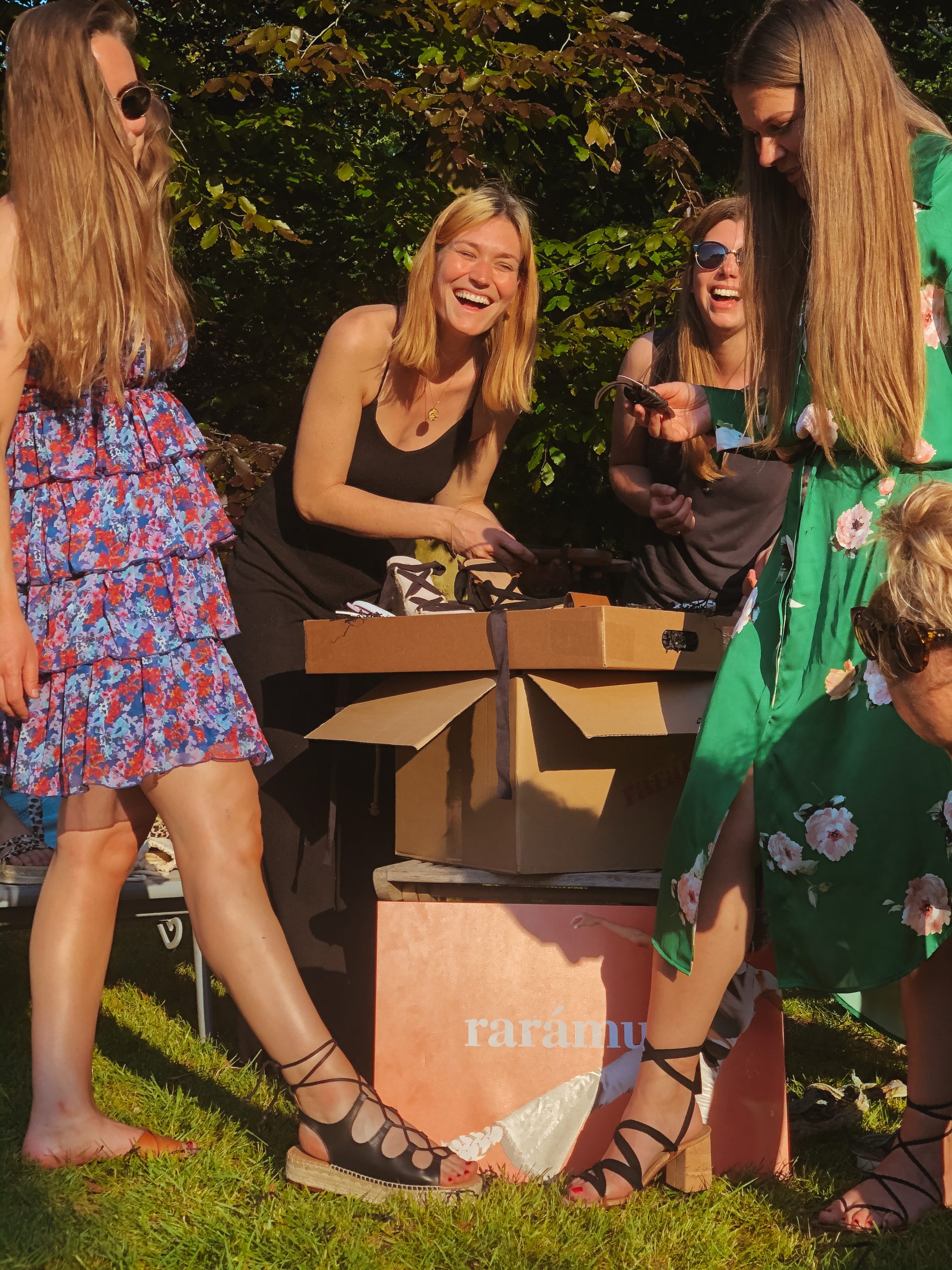 “Group of women enjoying a sunny outdoor gathering while unboxing Rarámuri sandals, wearing colorful summer dresses and black laced-up Rarámuri sandals.”